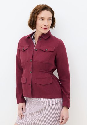 Mujer con cabello corto y castaño que lleva una chaqueta burdeos abotonada con bolsillos delanteros sobre una falda a rayas, mirando hacia su derecha.