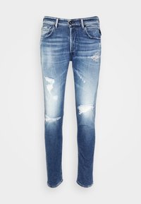 Jeans ajustados de mezclilla azul con un lavado desvaído y parches desgastados en los muslos y las rodillas, con bolsillos delanteros y presillas para cinturón.