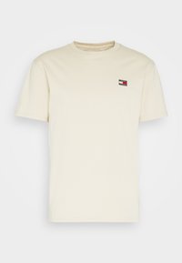 Krémovo zbarvené tričko s krátkým rukávem a malou nášivkou loga Tommy Jeans na levé straně hrudníku, umístěné na čistě bílém pozadí.