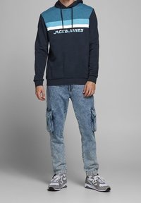 Navy collegehuppari teal- ja valkoisin raidoittein, logolla varustettuna. Yhdistetty vaaleansinisiin cargo-housuihin ja harmaisiin lenkkareihin.