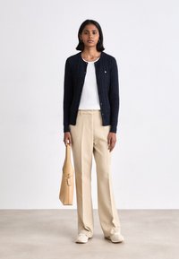 Donna che sta in piedi e tiene una borsa beige, indossa un cardigan blu scuro abbottonato, un top bianco, pantaloni beige e scarpe da ginnastica bianche su uno sfondo bianco semplice.