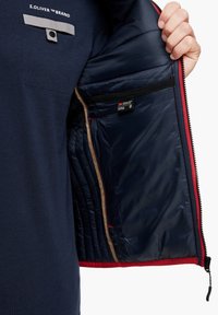 Veste isolante bleu marine avec des accents rouges ; dispose d'une poche zippée et d'une étiquette de marque à l'intérieur ; doublure intérieure en tissu lisse et brillant.