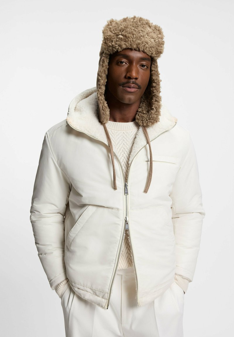 Witte gewatteerde jas met een capuchon gevoerd met nepbont, voorzien van een ritssluiting, zijzakken en gebreide details op de trui eronder.