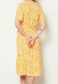 Abito floreale giallo con maniche corte, vita aderente e orlo svasato. Presenta fiori bianchi e dettagli rossi, realizzato in tessuto leggero.