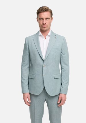 Man standing wearing a light gray textured suit with a white dress shirt, buttoned jacket, and matching trousers against a plain background.