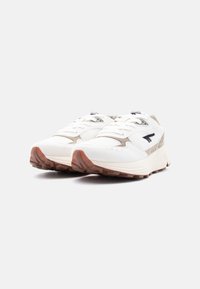 Ζευγάρι λευκά και μπεζ αθλητικά παπούτσια με καουτσούκ σόλες και μαύρο λογότυπο, πλάγια μπροστινή όψη σε ουδέτερο φόντο.