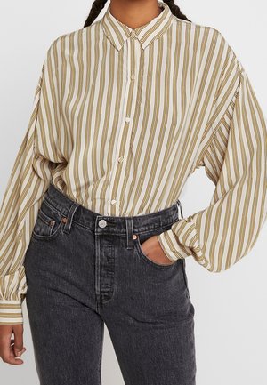 Kvinde iført en løs beige skjorte med lodrette brune striber, stoppet ind i højtaljede sorte denim-jeans, hånd i lommen.