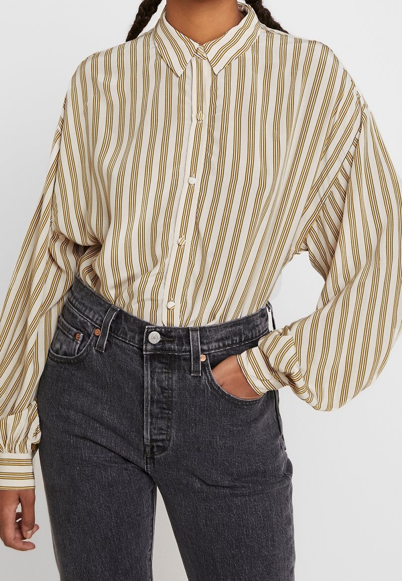 Donna che indossa una camicia beige ampia con strisce marroni verticali infilata in jeans di denim neri a vita alta, con una mano in tasca.