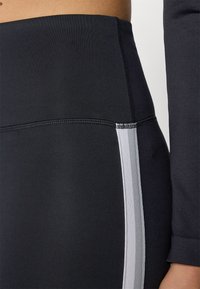 Hoge zwarte leggings met een gladde textuur en een grijze zijstreep, met stevige stiksels langs de tailleband en naden.
