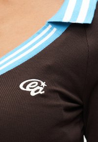 Καφέ μπλούζα με ραβδώσεις που διαθέτει γιακά με ουρανό μπλε και λευκές ρίγες, με λευκό κεντημένο λογότυπο στο στή θος.