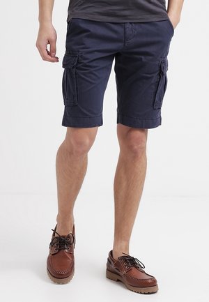 Homme portant un short cargo bleu marine et des chaussures en cuir marron à lacets, debout les mains dans les poches contre un fond uni.