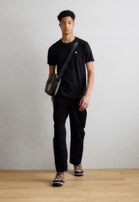 Crna majica, crne hlače i višebojne tenisice. Torba preko tijela u maslinasto zelenoj boji. Model stoji na drvenom podu nasuprot neutralnom zidu.