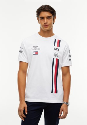 Ung mand iført en hvid T-shirt med røde, marineblå og sorte lodrette striber og flere motorsportslogoer på en hvid baggrund.