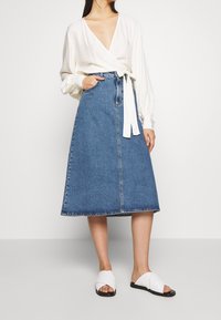 Bílá halenka s dlouhým rukávem a lehkým vzorem, spárovaná s modrou džínovou sukní ve tvaru A a bílými sandály s křížovými popruhy.