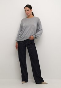 Jasnoszary, oversize sweter z okrągłym dekoltem i elastycznymi mankietami, zestawiony z ciemnoniebieskimi spodniami z szerokimi nogawkami i beżowymi butami na szpic.