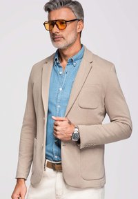 Homme aux cheveux gris et à la barbe portant des lunettes teintées orange, un blazer beige, une chemise en denim bleu, un pantalon blanc et une montre-bracelet, debout devant un fond neutre.