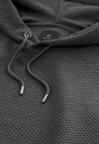 Mørkegrå hættetrøje med textured, quiltet stof. Har snore med metalspidser og et label, der viser "NEXT."