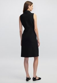 Schwarzes ärmelloses Kleid mit Kragen, A-Linie-Form, tailliert, glattes Material, knielang, kombiniert mit schwarzen flachen Schuhen.