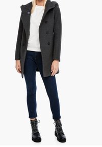 Grijze wollen blend jas met een capuchon, dubbele knoopsluiting en zwarte knopen; gecombineerd met een witte top, donkere skinny jeans en zwarte veterschoenen.