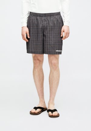 Mann trägt schwarz-weiß karierte Shorts, weißes Langarmshirt und schwarze Flip-Flops und steht vor einem weißen Hintergrund.