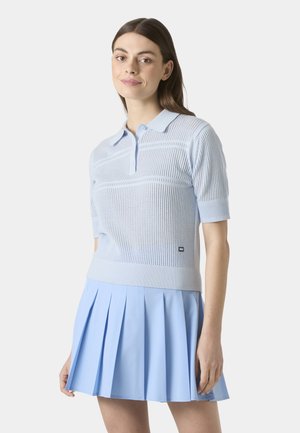 Mladá žena v světle modré lehké pletené polo košili s krátkými rukávy a plisované sukni, stojící před jednobarevným bílým pozadím.