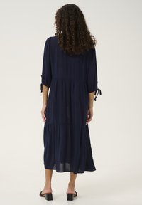 Vestido azul marino, de longitud hasta el tobillo, con mangas de tres cuartos, volantes fruncidos y detalles de lazo en las mangas. Tejido suave con un ajuste relajado.