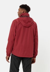 Červená voděodolná bunda s kapucí, která má elastické manžety a elegantní, minimalistický design. Nosená přes černé kalhoty.