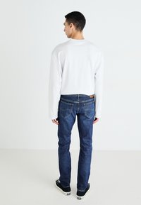 White long-sleeve shirt, blue denim jeans, and black sneakers. Jeans feature back pockets and a leather patch on the waistband.