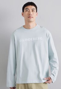 Hele sinine pika varrukaga särk ümmarguse kaelusega, mille esiküljel on julge valge kiri "J.LINDEBERG". Sujuv kangas, mugav lõige.