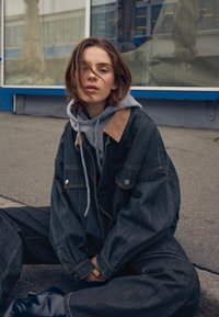 Giacca in denim con colletto in velluto, indossata sopra un hoodie grigio. Lavaggio scuro con vestibilità over, abbinata a pantaloni in denim larghi e stivali neri lucidi.
