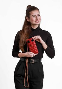 Červená prošívaná pouzdro na telefon se zlatým knoflíkem, odnímatelným řetízkovým popruhem a elegantním designem, které drží osoba v černém roláku a kalhotách s vysokým pasem.