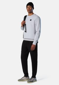 Felpa grigio chiaro con un piccolo logo, abbinata a pantaloni neri. Accessori includono un berretto nero e uno zaino. Le sneakers presentano accenti grigi e verdi.