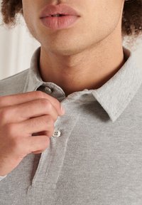 Graues Polohemd mit Kragen und Knopfleiste. Der Stoff wirkt weich und strukturiert, mit einer Nahaufnahme einer Hand, die den Kragen anpasst.