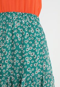 Jupe florale avec un fond bleu-vert, dotée d'une taille élastique et de marguerites blanches éparses avec un centre noir. Un ourlet à plusieurs niveaux ajoute de la texture.