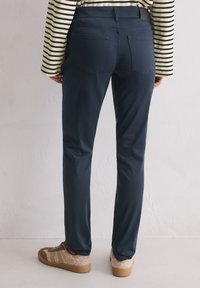 Marineblauwe, fitted broek met een gladde textuur, twee achterzakken en een middelhoge taille, gedragen met bruine sneakers en een gestreept shirt.