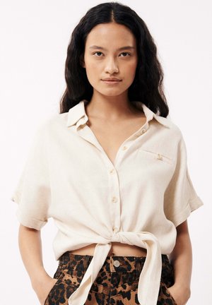 Chemise à manches courtes et à boutons en tissu beige clair avec une finition texturée. Présente une poche avant et un ourlet noué. Associée à un short à imprimé léopard.