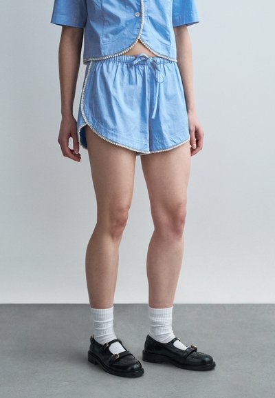 Personne portant une tenue deux pièces bleu clair avec bordure de perles, chaussettes blanches montantes et chaussures brogues en cuir noir, debout sur un sol gris.