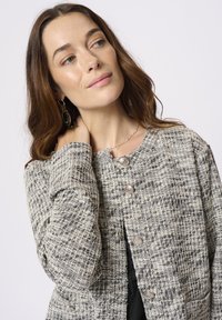 Texturovaná, tkaná bunda v černé a krémové barvě. Má kulatý výstřih, zapínání na knoflíky a dvě kapsy. Tkanina vypadá teple a útulně.