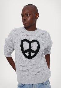 Γκρι πλεκτό πουλόβερ με υφή, διακοσμημένο με ένα μαύρο σύμβολο ειρήνης ραμμένο στο μπροστινό μέρος. Κοντά μανίκια και στρογγυλή λαιμόκοψη.