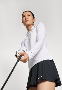 Bílé sportovní triko s dlouhým rukávem a límcem, v kombinaci s černou mnohovrstvou sukní. V jedné ruce drží golfovou hůl, na rukou má bílé rukavice.
