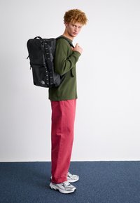 Svart ryggsäck med flera fack och justerbara remmar, bärs av en modell i en grön tröja och röda byxor, mot en neutral bakgrund.