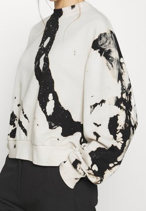 White sweatshirt with black abstract paint splatter design, long puffed sleeves, and ribbed cuffs worn with black pants.