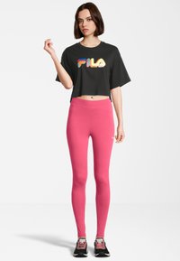 T-shirt cropped nera con logo "FILA" colorato, abbinata a leggings rosa. Il modello indossa sneakers nere con dettagli rosa. Tessuto morbido.
