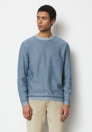 Blauer gestrickter Pullover mit Rundhalsausschnitt, der ein strukturiertes Muster und gerippte Bündchen aufweist. Kombiniert mit beigen Hosen.