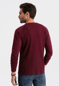 Pitkähihaine burguinivärinen ribbipaita, jossa pyöreä kaula-aukko ja raglan-hihat, yhdistettynä farkkuihin. Tekstuuriltaan pehmeä ja istuva siluetti.