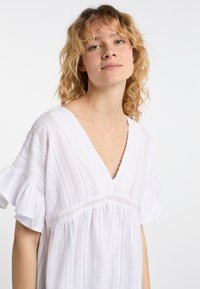 Hvid bomuldsbluse med V-udskæring, korte volangærmer og struktureret stribet mønster. Har blonde detaljer langs udskæringen.