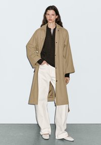 Femme debout, la main dans la poche de son pantalon blanc, portant un trench-coat beige, un pull noir et des chaussures blanches, devant un fond clair.