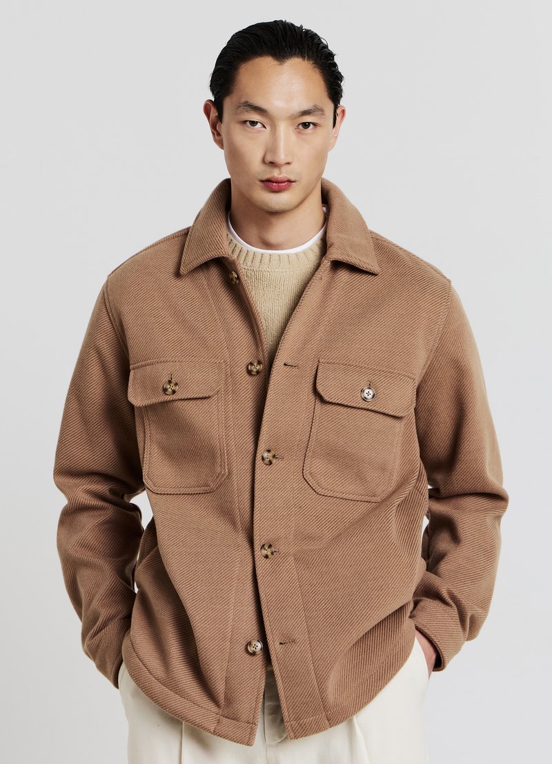 Giovane uomo con capelli scuri indossa una giacca beige con bottoni sopra a un maglione leggero lavorato a maglia e pantaloni bianchi su uno sfondo chiaro e uniforme.
