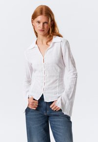 Camicia bianca con bottoni, collo a camicia, tessuto strutturato e maniche svasate, indossata sopra jeans blu. Presenta sottili righe verticali.