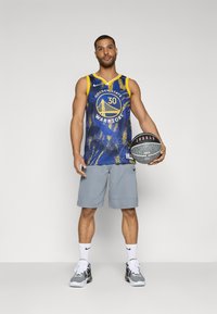 Golden State Warriors basketballdrakt i blå og gul med mønsterdesign, grå shorts, holder en svart og hvit basketball.